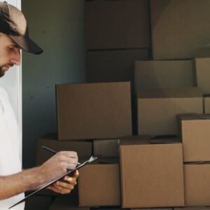 Deliveryman writing on clipboard next to stacked cardboard boxes in a van.